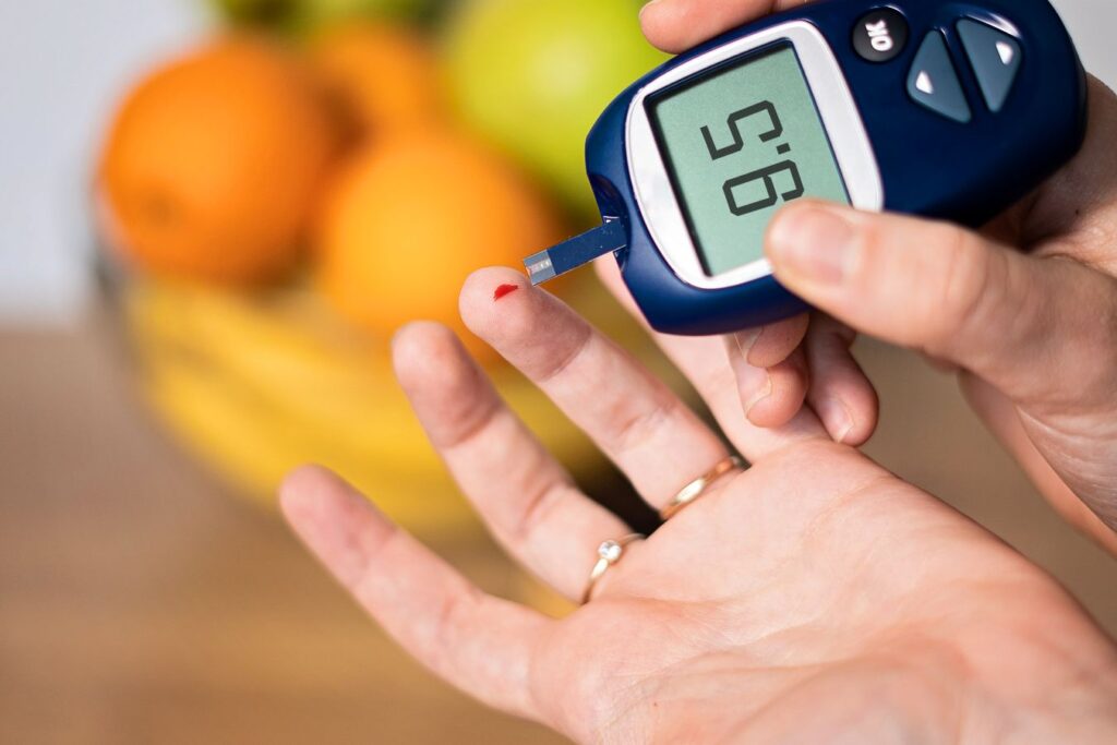 Pessoa medindo a glicose no sangue com glicosímetro, mostrando resultado de 95 mg/dL, com frutas desfocadas ao fundo.