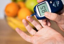 Pessoa medindo a glicose no sangue com glicosímetro, mostrando resultado de 95 mg/dL, com frutas desfocadas ao fundo.