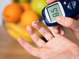 Pessoa medindo a glicose no sangue com glicosímetro, mostrando resultado de 95 mg/dL, com frutas desfocadas ao fundo.
