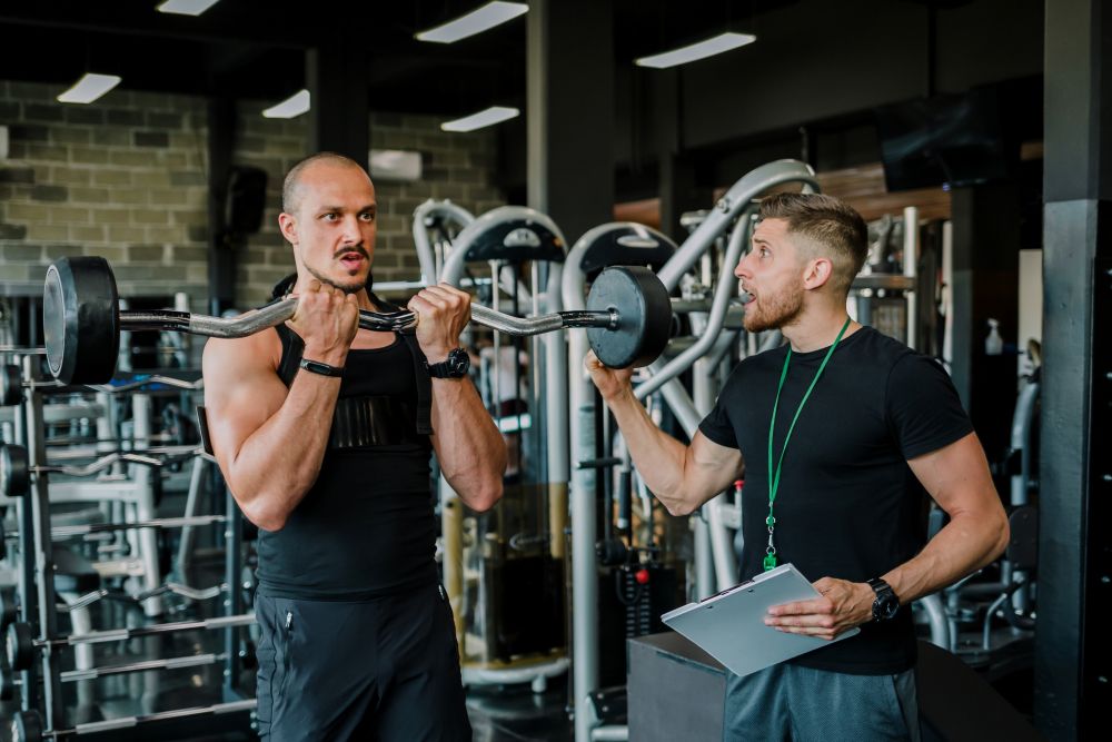Homem treinando bíceps com barra enquanto personal trainer orienta e acompanha em academia.