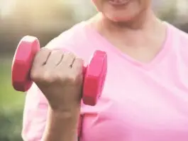 Pessoa usando camiseta rosa segura um halter rosa durante exercício ao ar livre, em imagem associada à atividade física e à prevenção do câncer de mama.