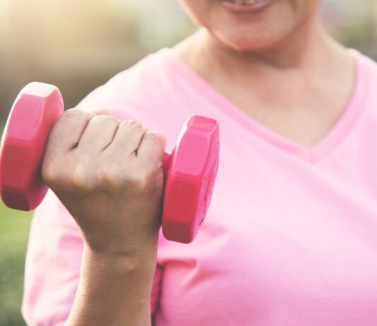 Pessoa usando camiseta rosa segura um halter rosa durante exercício ao ar livre, em imagem associada à atividade física e à prevenção do câncer de mama.