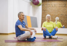 Casal de idosos praticando ginástica no solo em academia, sentados em posição de meditação sobre tapetes de yoga.