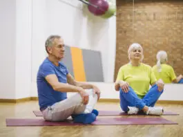 Casal de idosos praticando ginástica no solo em academia, sentados em posição de meditação sobre tapetes de yoga.