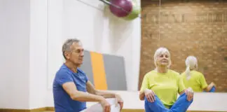 Casal de idosos praticando ginástica no solo em academia, sentados em posição de meditação sobre tapetes de yoga.