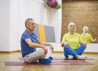 Ginástica para idosos: mais saúde, equilíbrio e autonomia Casal de idosos praticando ginástica no solo em academia, sentados em posição de meditação sobre tapetes de yoga.
