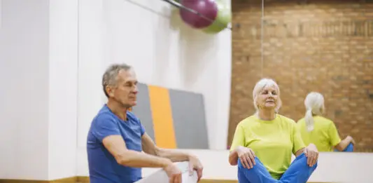 Ginástica para idosos: mais saúde, equilíbrio e autonomia Casal de idosos praticando ginástica no solo em academia, sentados em posição de meditação sobre tapetes de yoga.