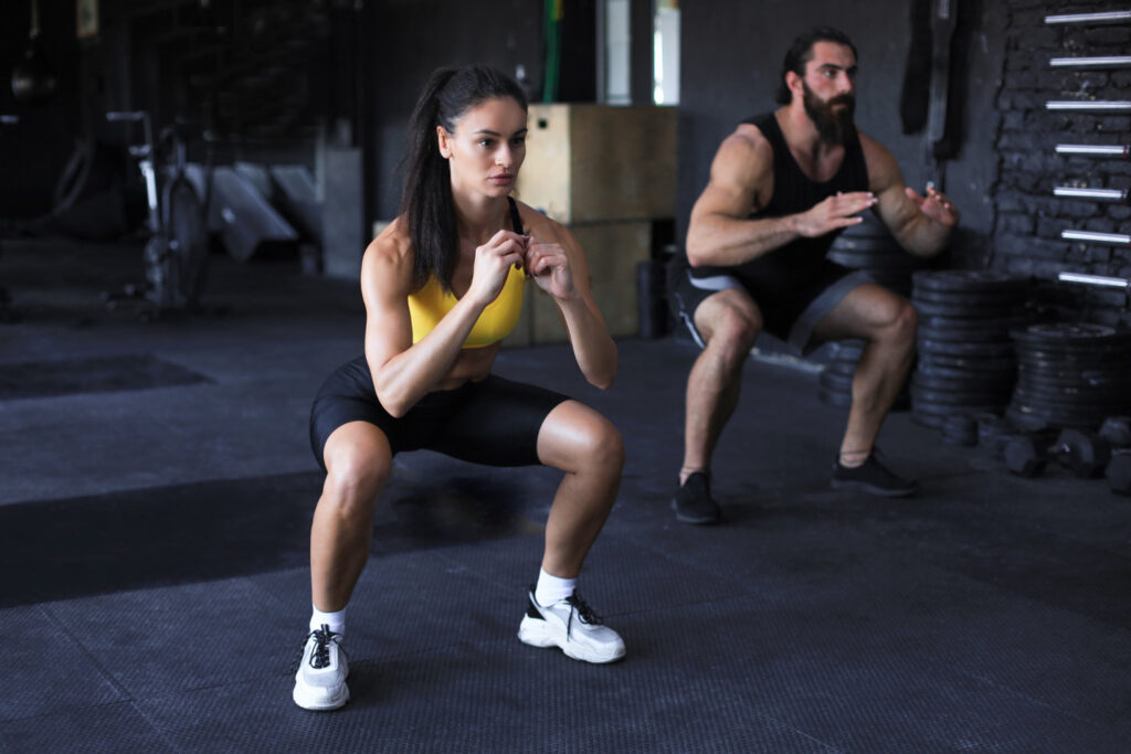 Casal fitness executando agachamentos em academia moderna, demonstrando um dos exercícios mais eficazes para quem busca como engrossar as pernas.