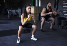Casal fitness executando agachamentos em academia moderna, demonstrando um dos exercícios mais eficazes para quem busca como engrossar as pernas.