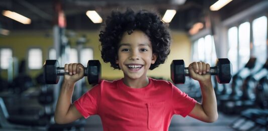 Pequeno atleta simula treino de musculação para crianças e adolescentes, segurando halteres com as mãos.