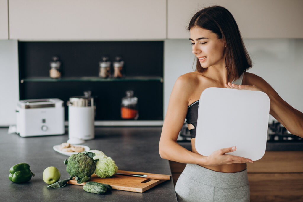 Mulher segurando balança na cozinha com alimentos saudáveis ao lado.