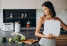Mulher segurando balança na cozinha com alimentos saudáveis ao lado.
