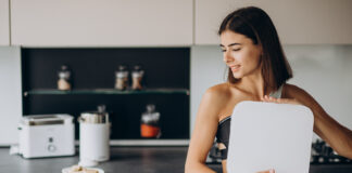 Mulher segurando balança na cozinha com alimentos saudáveis ao lado.