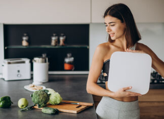 Mulher segurando balança na cozinha com alimentos saudáveis ao lado.