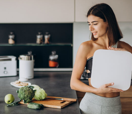 Mulher segurando balança na cozinha com alimentos saudáveis ao lado.