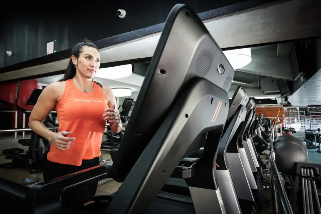 Mulher corre em uma esteira dentro de uma academia, usando regata esportiva laranja e calça preta, com outros equipamentos de cardio ao fundo.