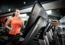 Mulher corre em uma esteira dentro de uma academia, usando regata esportiva laranja e calça preta, com outros equipamentos de cardio ao fundo.