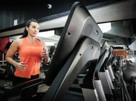 Mulher corre em uma esteira dentro de uma academia, usando regata esportiva laranja e calça preta, com outros equipamentos de cardio ao fundo.