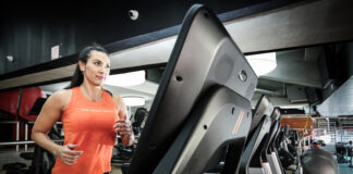 HIIT na esteira: descubra se esse treino combina com seu objetivo Mulher corre em uma esteira dentro de uma academia, usando regata esportiva laranja e calça preta, com outros equipamentos de cardio ao fundo.