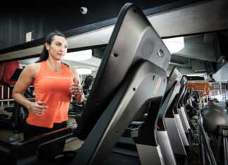 Mulher corre em uma esteira dentro de uma academia, usando regata esportiva laranja e calça preta, com outros equipamentos de cardio ao fundo.