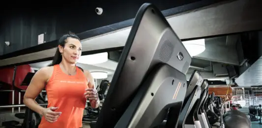 Mulher corre em uma esteira dentro de uma academia, usando regata esportiva laranja e calça preta, com outros equipamentos de cardio ao fundo.