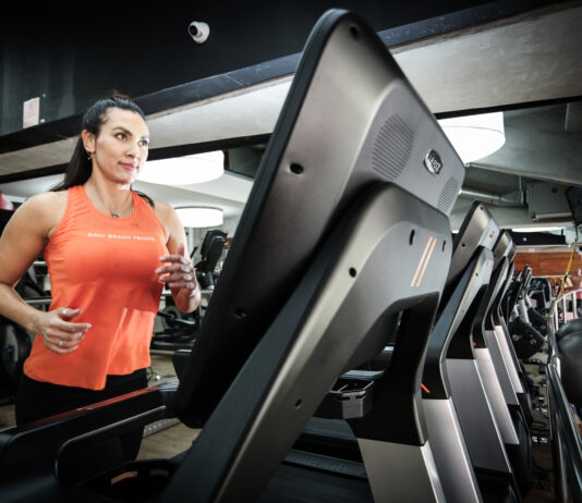 Mulher corre em uma esteira dentro de uma academia, usando regata esportiva laranja e calça preta, com outros equipamentos de cardio ao fundo.