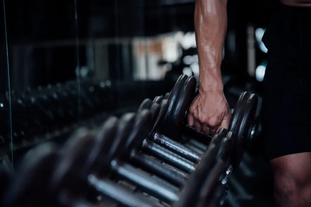 Pessoa segurando um halter em academia durante treino de musculação, com foco no braço e nos pesos em ambiente escuro e moderno.