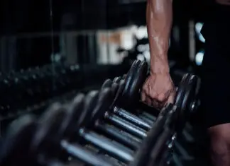 Pessoa segurando um halter em academia durante treino de musculação, com foco no braço e nos pesos em ambiente escuro e moderno.