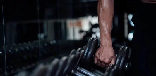 Pessoa segurando um halter em academia durante treino de musculação, com foco no braço e nos pesos em ambiente escuro e moderno.