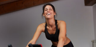 Mulher sorri enquanto pedala em uma bicicleta ergométrica durante treino cardiovascular, ilustrando a prática de exercícios aeróbicos para emagrecimento e melhora do condicionamento físico.