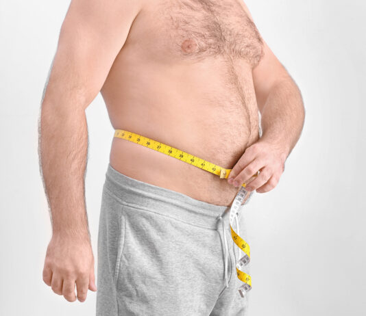 Homem adulto mede a circunferência da barriga com fita métrica, evidenciando gordura abdominal.