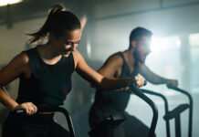 Mulher e homem realizando exercícios aeróbicos em equipamentos de academia, em ambiente interno de treinamento.