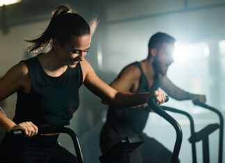 Mulher e homem realizando exercícios aeróbicos em equipamentos de academia, em ambiente interno de treinamento.