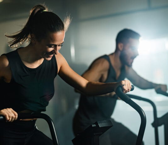 Mulher e homem realizando exercícios aeróbicos em equipamentos de academia, em ambiente interno de treinamento.