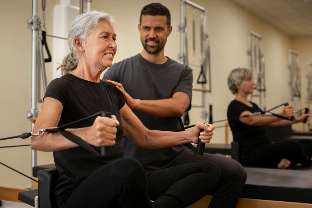 Idosa realiza exercício de pilates em aparelho acompanhada por instrutor.