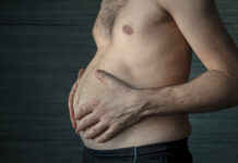 Homem segura a própria barriga em close lateral, destacando o acúmulo de gordura abdominal em ambiente escuro e neutro.