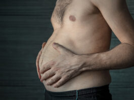 Nem toda barriga é igual: o perigo invisível da gordura visceral Homem segura a própria barriga em close lateral, destacando o acúmulo de gordura abdominal em ambiente escuro e neutro.