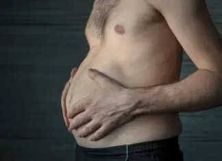 Homem segura a própria barriga em close lateral, destacando o acúmulo de gordura abdominal em ambiente escuro e neutro.
