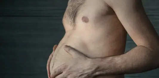 Nem toda barriga é igual: o perigo invisível da gordura visceral Homem segura a própria barriga em close lateral, destacando o acúmulo de gordura abdominal em ambiente escuro e neutro.