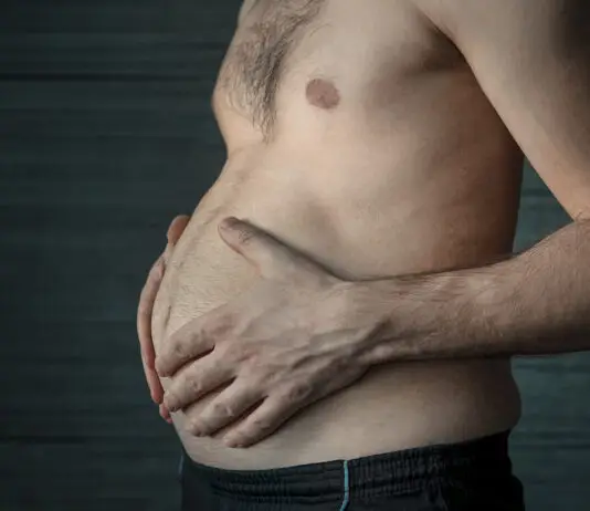 Homem segura a própria barriga em close lateral, destacando o acúmulo de gordura abdominal em ambiente escuro e neutro.