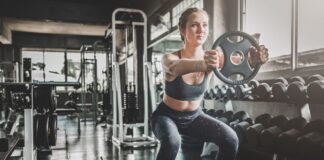 Mulher jovem com roupa fitness faz agachamento livre segurando anilha de peso em ambiente de academia