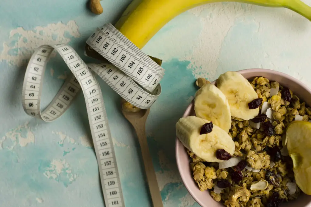 Fita métrica enrolada em uma banana ao lado de tigela com granola e frutas, representando alimentação saudável e déficit calórico.