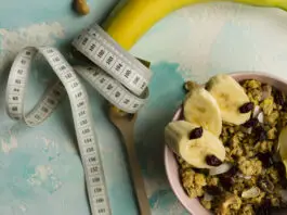 Fita métrica enrolada em uma banana ao lado de tigela com granola e frutas, representando alimentação saudável e déficit calórico.
