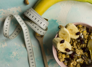 Déficit calórico não é passar fome, e esse erro atrapalha muita gente Fita métrica enrolada em uma banana ao lado de tigela com granola e frutas, representando alimentação saudável e déficit calórico.