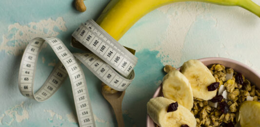 Déficit calórico não é passar fome, e esse erro atrapalha muita gente Fita métrica enrolada em uma banana ao lado de tigela com granola e frutas, representando alimentação saudável e déficit calórico.