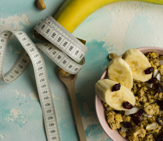 Déficit calórico não é passar fome, e esse erro atrapalha muita gente Fita métrica enrolada em uma banana ao lado de tigela com granola e frutas, representando alimentação saudável e déficit calórico.