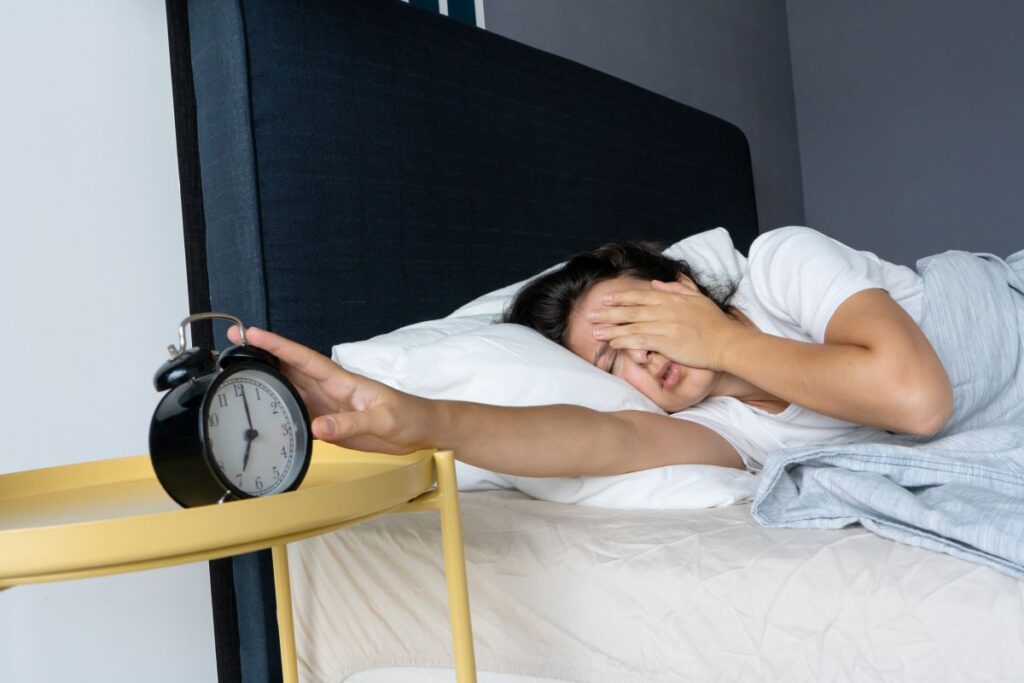 Deitada na cama e com sono, mulher desliga o despertador. Acordar cansado é comum para muitas pessoas.