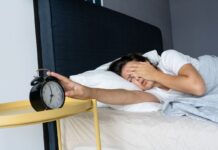 Deitada na cama e com sono, mulher desliga o despertador. Acordar cansado é comum para muitas pessoas.