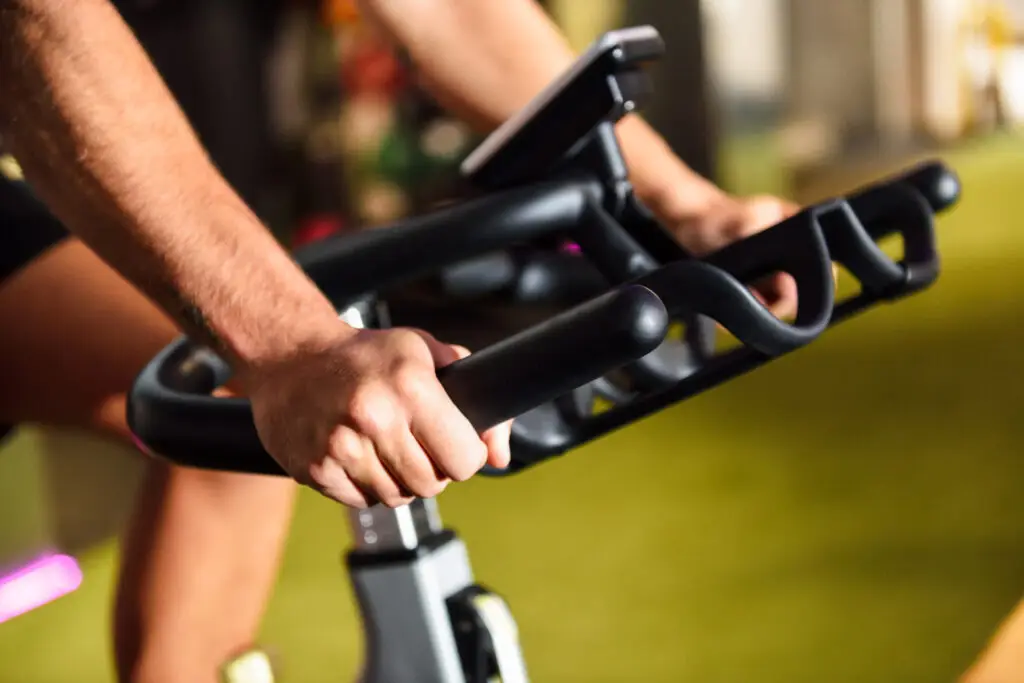 Close-up das mãos de uma pessoa utilizando uma bicicleta ergométrica em uma academia. A imagem destaca a pegada firme no guidão e o painel do equipamento ao fundo, representando um exercício cardiovascular de baixo impacto.