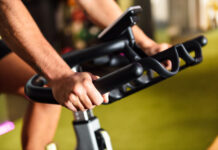 Close-up das mãos de uma pessoa utilizando uma bicicleta ergométrica em uma academia. A imagem destaca a pegada firme no guidão e o painel do equipamento ao fundo, representando um exercício cardiovascular de baixo impacto.